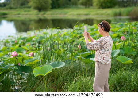 Adult woman takes pictures on a phone camera of a beautiful landscape with a lake of lotuses. Travel concept.