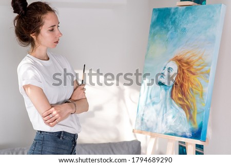 beautiful woman holding paintbrush, happy looking enjoying picture, inspired drawing picture, girls with orange hair. Home workshop, enjoyment creativity inspiration. Art painting easel.