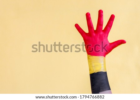 A little girl's child's hand painted in the Belgian flag shows hello to the right and with space for text on the left on a yellow background, close-up side view.