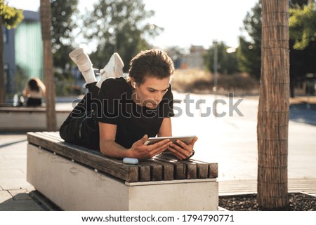 Young man watching movie on digital tablet while lying on bench at college campus. High quality photo