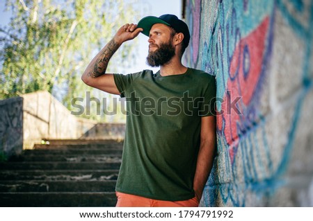 City portrait of handsome hipster guy with beard wearing a blank green military t-shirt and black cap. Empty space for your logo or design. Mockup for print.