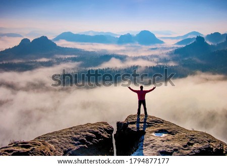 Small silhouette of hiker man enjoying beautiful sunrise in morning mountains. Traveler with raised hands standing on mountain with white fog below. Royalty-Free Stock Photo #1794877177