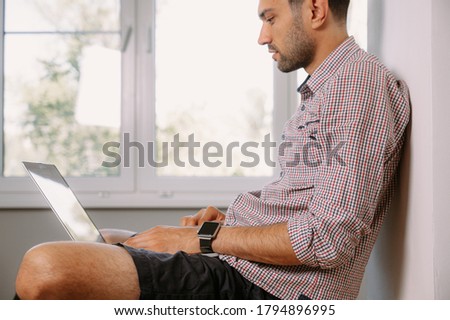 A young man in a shirt sits on the windowsill and works on a laptop at home.Young man working on laptop at home