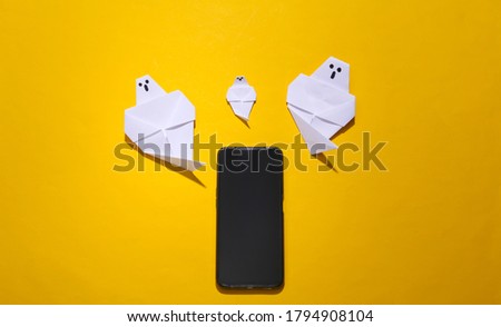 Origami ghosts and smartphone on yellow bright background. Top view