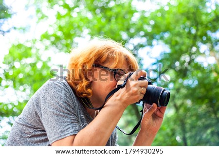 Redhead woman photographing on city park. Tourist hold, looking in camera and taking pictures. Professional photographer taking camera outdoors in nature