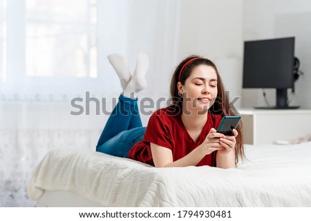 A young woman is lying on the bed on her stomach and typing something in her smartphone. Copy space. White interior of the room in the background. Concept of modern technologies and communications