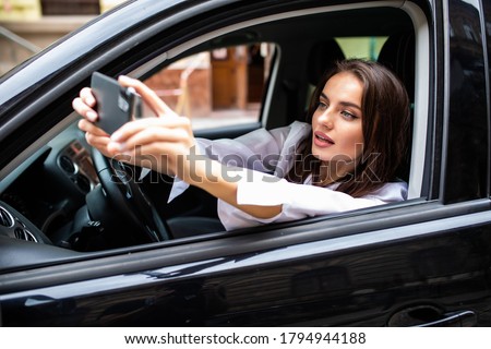 Happy young woman holding mobile phone and taking photos while driving a car. Smiling girl taking selfie picture with smart phone camera outdoors in car.