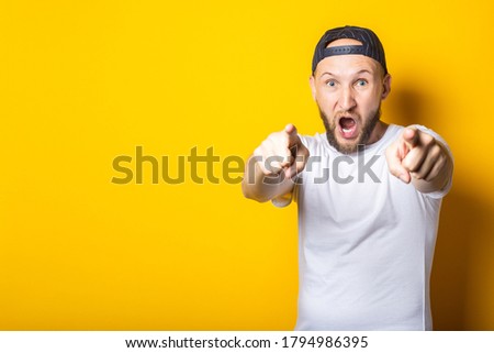 Bearded young man in white t-shirt shouting and pointing fingers at camera on yellow background.