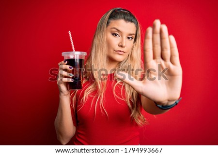 Young beautiful blonde woman drinking cola fizzy beverage using straw over red background with open hand doing stop sign with serious and confident expression, defense gesture