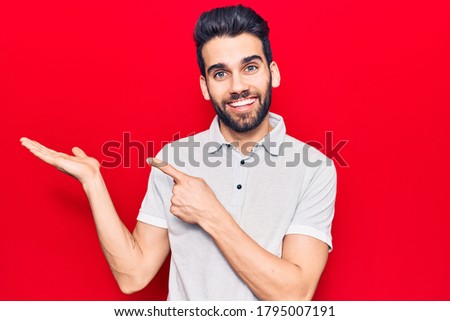 Young handsome man with beard wearing casual polo amazed and smiling to the camera while presenting with hand and pointing with finger. 