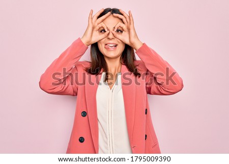 Young beautiful brunette businesswoman wearing elegant jacket over isolated pink background doing ok gesture like binoculars sticking tongue out, eyes looking through fingers. Crazy expression.