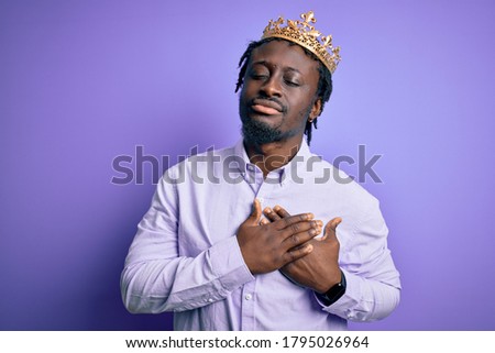 Young african american man wearing golden crown of king over isolated purple background smiling with hands on chest with closed eyes and grateful gesture on face. Health concept.