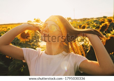 Cheerful joyful young woman with closed eyes posing on camera. Stand alone in middle of sunflower's field. Sunny morning or evening. Natural picture or real people. Touching hair and enjoy summertime