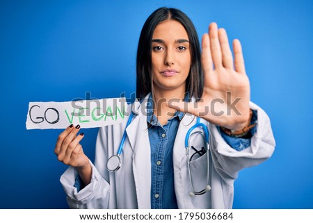 Young beautiful brunette doctor woman wearing coat holding paper with go vegan message with open hand doing stop sign with serious and confident expression, defense gesture