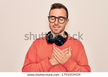 Young handsome man listening to music using headphones over isolated white background smiling with hands on chest with closed eyes and grateful gesture on face. Health concept.
