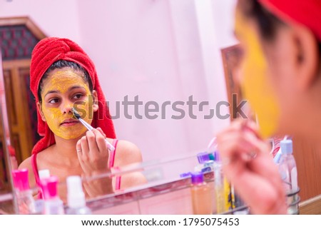 Skin care - Beautiful girl applying Gram flour (Besan) turmeric yellow face mask on face through brush. she is wearing red towel on head and standing front of the mirror at bathroom.
 Royalty-Free Stock Photo #1795075453
