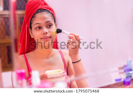 Skin care - Beautiful girl applying Gram flour (Besan) turmeric yellow face mask on face through brush. she is wearing red towel on head and standing front of the mirror at bathroom.
 Royalty-Free Stock Photo #1795075468