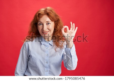 Smiling curly redhead woman showing Ok sign with her left hand on the red background