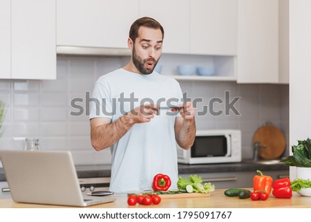 Shocked young bearded guy in white t-shirt taking pictures on mobile phone preparing vegetable salad cooking food in light kitchen at home. Dieting healthy lifestyle concept. 