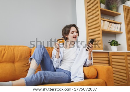 Cheerful young woman in casual clothes sit on couch spending time in living room at home. Rest relax good mood leisure lifestyle concept. Mock up copy space. Using mobile phone, hold credit bank card