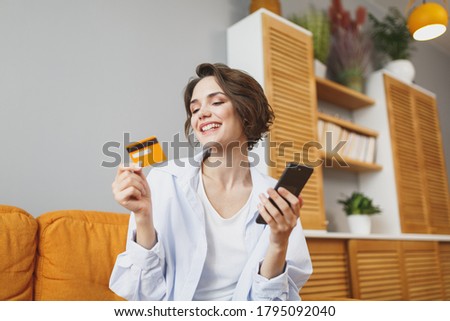 Beautiful young woman in casual clothes sit on couch spending time in living room at home. Rest relax good mood leisure lifestyle concept. Mock up copy space. Using mobile phone hold credit bank card