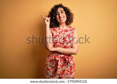 Young beautiful curly arab woman on vacation wearing summer floral dress and sunglasses with a big smile on face, pointing with hand and finger to the side looking at the camera.