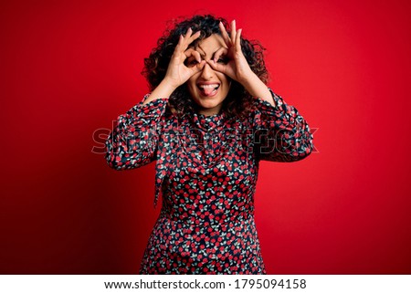 Young beautiful curly arab woman wearing casual floral dress standing over red background doing ok gesture like binoculars sticking tongue out, eyes looking through fingers. Crazy expression.
