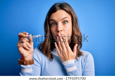 Young blonde girl holding dental orthodontic clear aligner over blue isolated background cover mouth with hand shocked with shame for mistake, expression of fear, scared in silence, secret concept