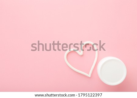 Heart shape created from cream. White jar on light pink table background. Pastel color. Care about face, hands, legs and body skin. Closeup. Empty place for text or logo.