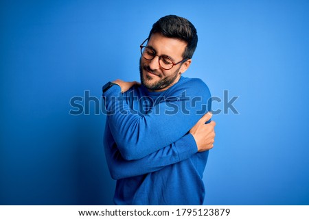 Young handsome man with beard wearing casual sweater and glasses over blue background Hugging oneself happy and positive, smiling confident. Self love and self care Royalty-Free Stock Photo #1795123879