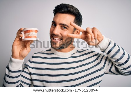 Young handsome man with beard holding plastic denture teeth over white background Doing peace symbol with fingers over face, smiling cheerful showing victory