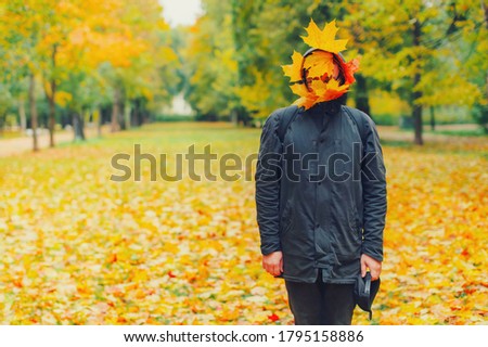 Strange hipster man in black parka jacket with fall leaves on face standing in autumn park. Funny trendy fashion male model creative portrait outdoor. Conceptual creepy guy in autumn weather street.