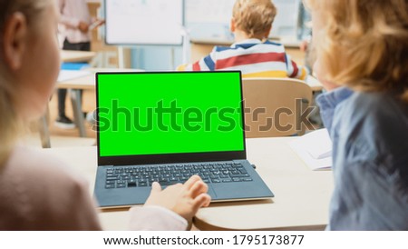 Elementary School Science Class: Over the Shoulder Little Boy and Girl Use Laptop with Green Screen Mock-up Template on a Display. Physics Teacher Explains Lesson to a Diverse Class full of Smart Kids