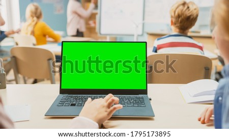 Elementary School Science Class: Little Girl Use Laptop with Green Screen Mock-up Template. In the Background School Class full of Diverse Smart Kids