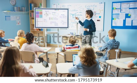 Elementary School Science Teacher Uses Interactive Digital Whiteboard to Show Classroom Full of Children how Software Programming works for Robotics. Science Class, Curious Kids Listening Attentively Royalty-Free Stock Photo #1795173919
