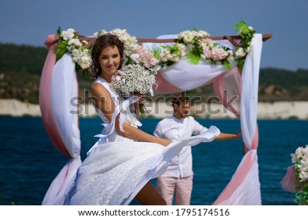 the bride and groom in white clothes with a bouquet of white flowers stand under an arch of flowers and fabric against the background of a blue lake and white sand