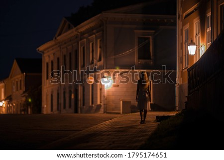 Young alone woman in white dress slowly walking on sidewalk under street lights at old town in dark summer night. Peaceful atmosphere. Spending time alone. City life. Back view.  Royalty-Free Stock Photo #1795174651