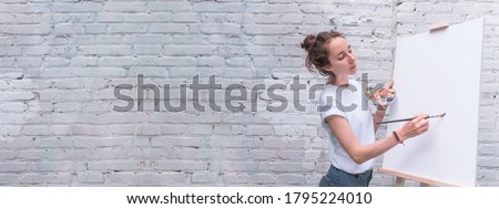 Beautiful woman artist, brick wall background, free space for copy of text, draws a picture, white T-shirt, brush in hand, palette for paints. White canvas, easel.