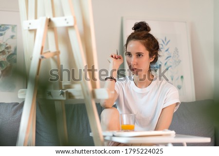 Beautiful woman artist in a white T-shirt, at home on couch, brush in hand, table with paints. Background, easel and wall with paintings. Looks at the painting.