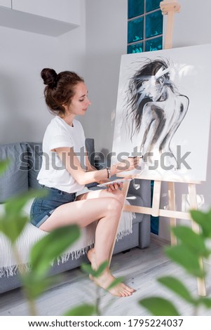 Woman artist, draws a silhouette of a girl with long hair on a white background, at home in the workshop, in her hand a brush. Background sofa, easel green flowers out of focus.