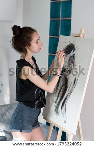 Woman artist, draws a silhouette of a girl on canvas, at home in workshop, in his hand a brush. Background, easel and wall with paintings.