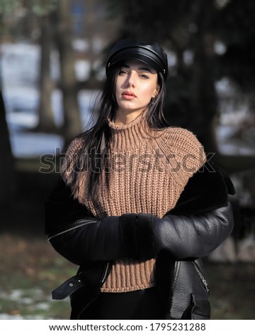 Young brunette woman in the leather pants sheepskin coat and black cappi posing at the park