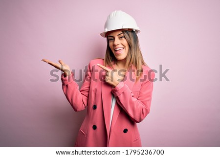 Young beautiful brunette architect woman wearing safety helmet over pink background amazed and smiling to the camera while presenting with hand and pointing with finger.
