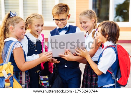 Education at school. Classmates in school uniforms standing in courtyard against the school building. The older boy holding laptop. Children looking to, smiling, laughing, showing each other pictures