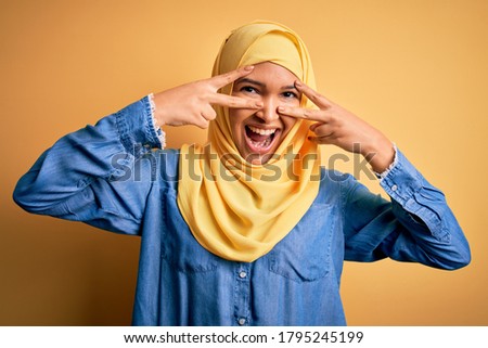 Young beautiful woman with curly hair wearing arab traditional hijab over yellow background Doing peace symbol with fingers over face, smiling cheerful showing victory