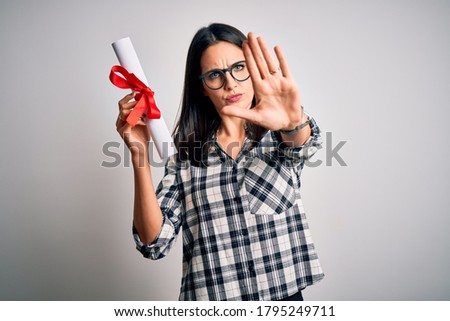 Young brunette student woman with blue eyes holding university diploma over isolated background with open hand doing stop sign with serious and confident expression, defense gesture