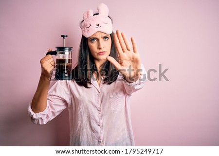 Young brunette woman with blue eyes wearing pajama making coffee with french coffeemaker with open hand doing stop sign with serious and confident expression, defense gesture