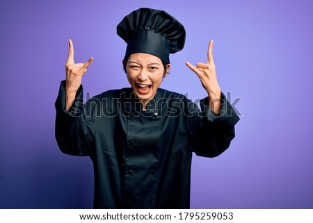 Young beautiful chinese chef woman wearing cooker uniform and hat over purple background shouting with crazy expression doing rock symbol with hands up. Music star. Heavy music concept.