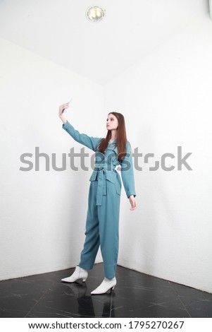European Belarusian girl is making a selfie in a navy blue summer spring suit in a room with white walls, studio portrait of an influencer podcasting.