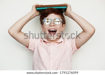 Young caucasian boy with ginger hair holding some book s on his head and smile at camera wearing eyeglasses